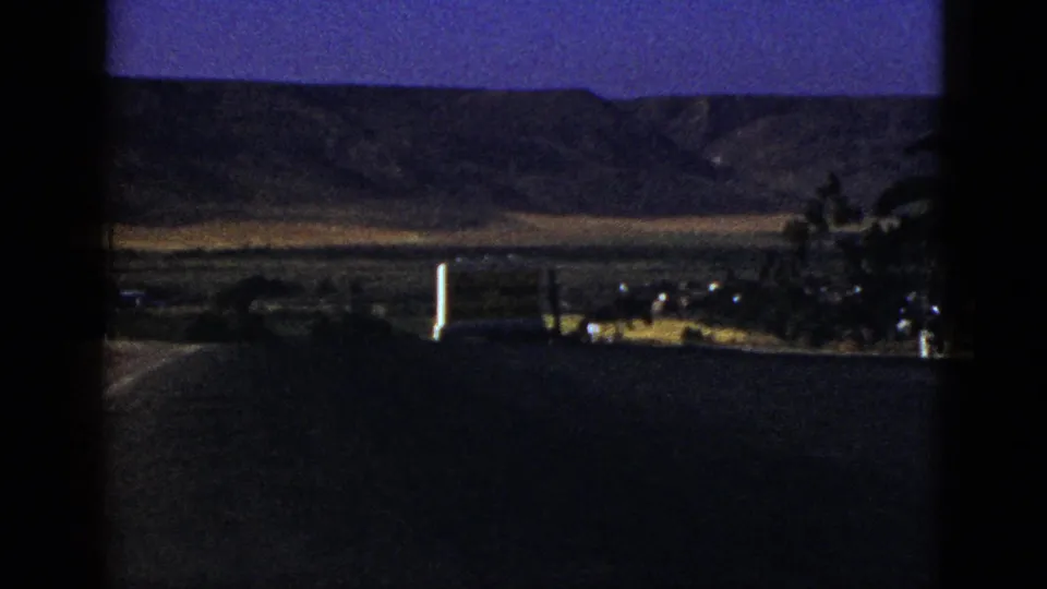 Dim 8mm frame looking across a paved road toward distant California hills, with signs and trees at the roadside.