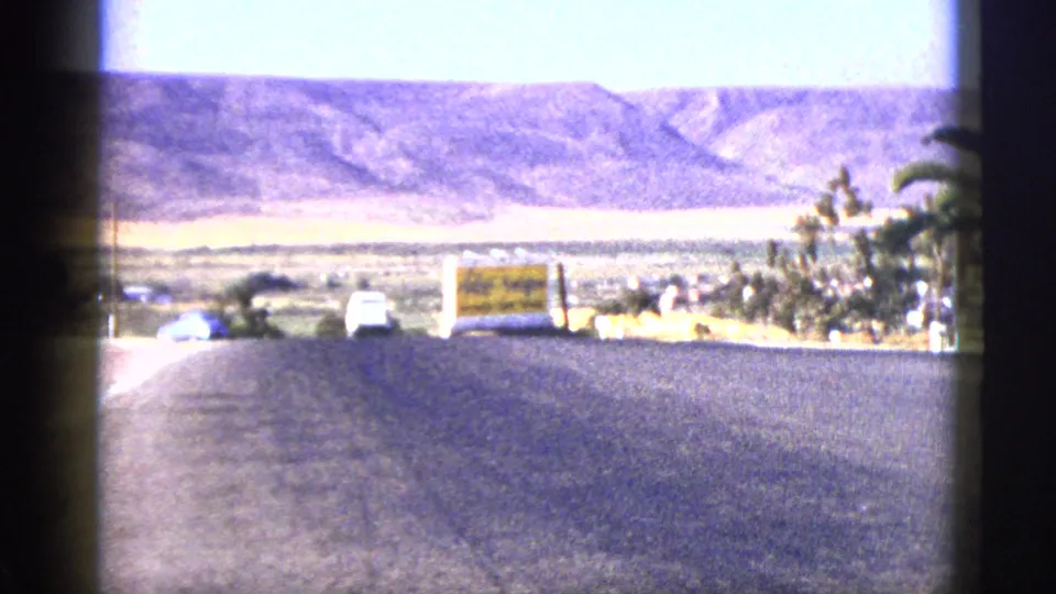 1953 California home-movie frame of a broad road descending toward hills, with cars and a yellow roadside sign.