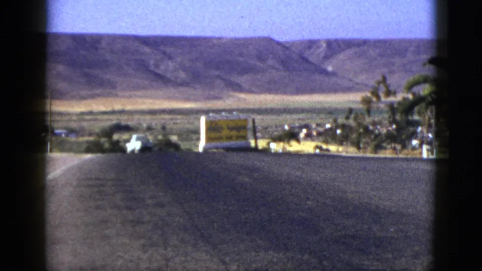 Grainy 8mm California road view with distant hills, palm trees, cars, and a yellow sign likely reading Tecate.
