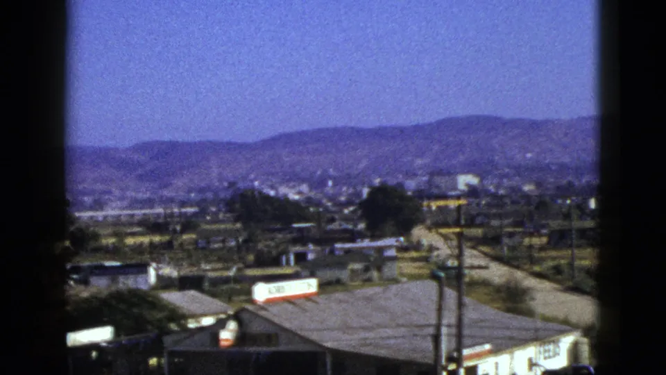 1953 California townscape frame showing rooftops, roads, utility poles, small signs, distant buildings, and mountains under a blue sky.