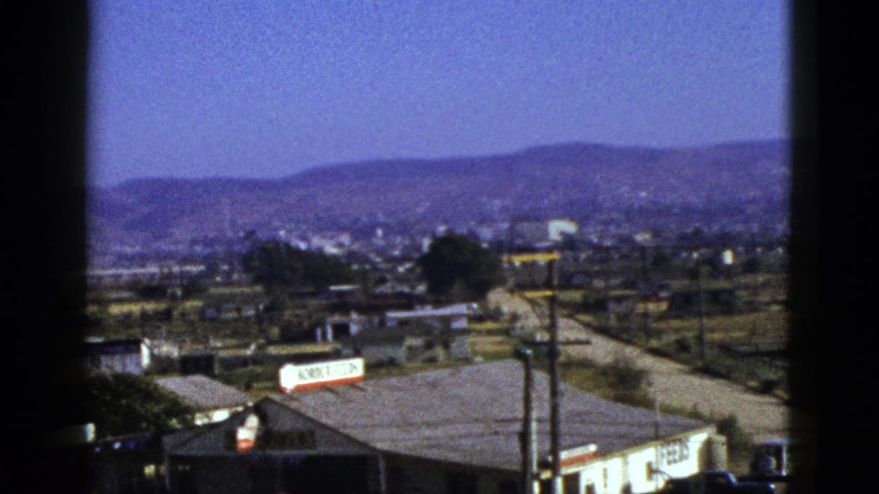 1953 California home-movie frame showing a foothill town edge through a window, with roadside buildings, utility poles, and a Border Feeds sign.
