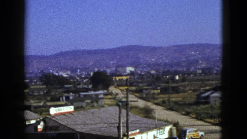 1953 California foothill town view through a window, showing roadside buildings, a receding road, utility poles, signage, and a yellow car.