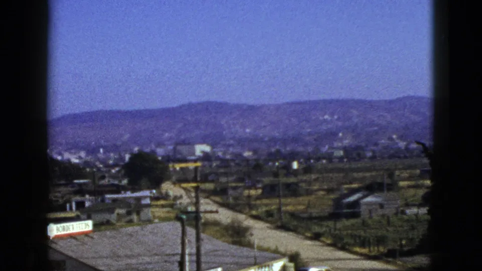 1953 California 8mm frame with rooftops, a roadside sign, utility poles, open lots, and distant foothills under a blue sky.