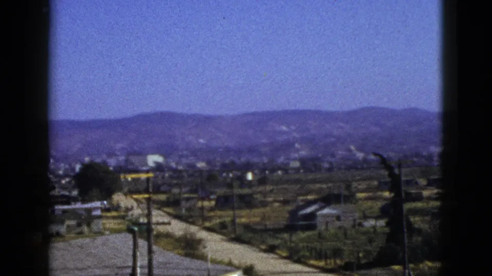 1953 California town-edge view through a window, with foothills, low buildings, roadside lots, a road, and utility poles in soft 8mm color.