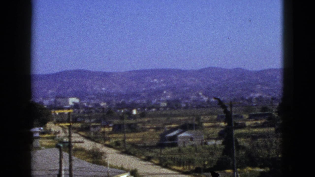 1953 California home-movie view of a valley town, pale road, utility poles, fields, and distant hills framed by dark window edges.