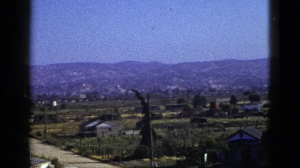 Mid-century 8mm view across a California valley, with a pale road, utility poles, low buildings, planted fields, and rolling hills.