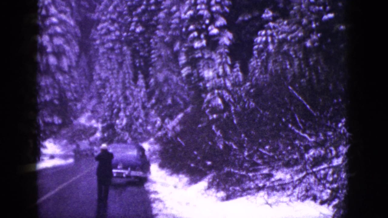 1953 California 8mm frame showing a snowy forest road, a person in dark clothing, and a parked car under evergreen trees.