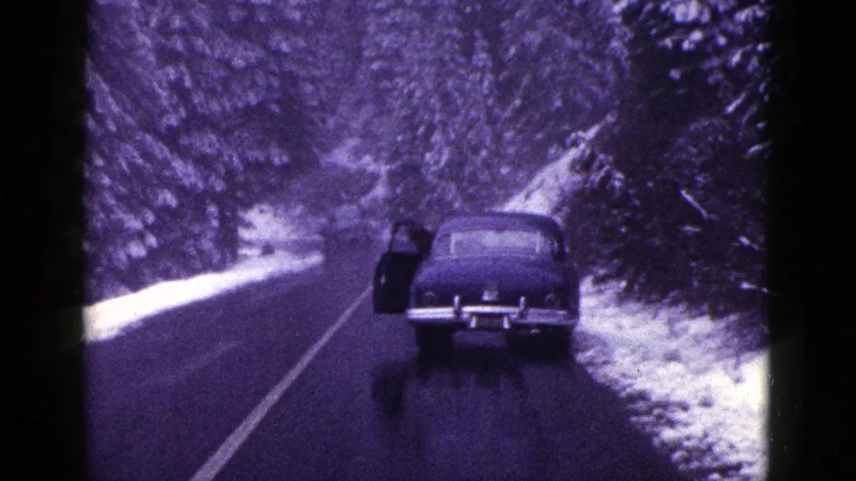 1953 California home-movie frame with a dark car parked on a wet snowy forest road, its door open.