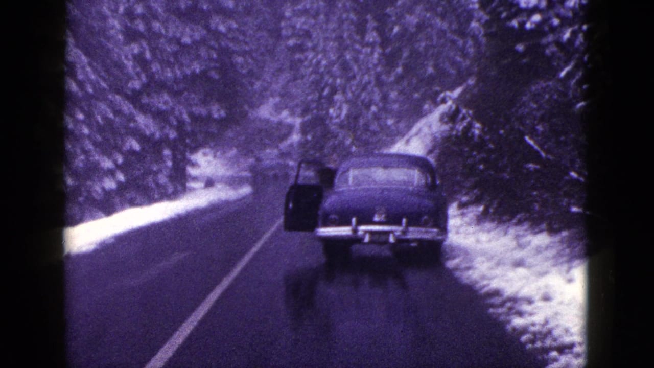 1953 California 8mm frame showing a dark sedan on a wet mountain road bordered by snow and evergreen trees.