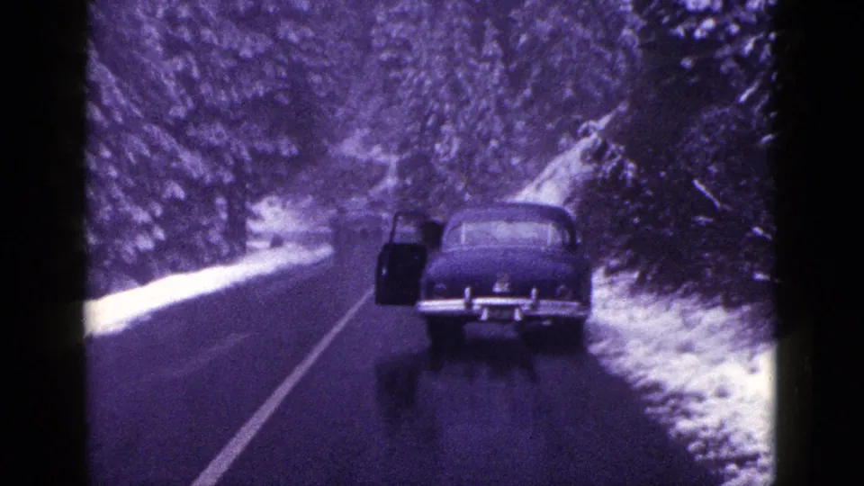 1953 California 8mm frame showing a dark sedan on a wet mountain road bordered by snow and evergreen trees.