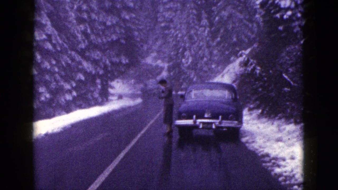 Man in a dark coat stands near a stopped sedan on a snowy road.