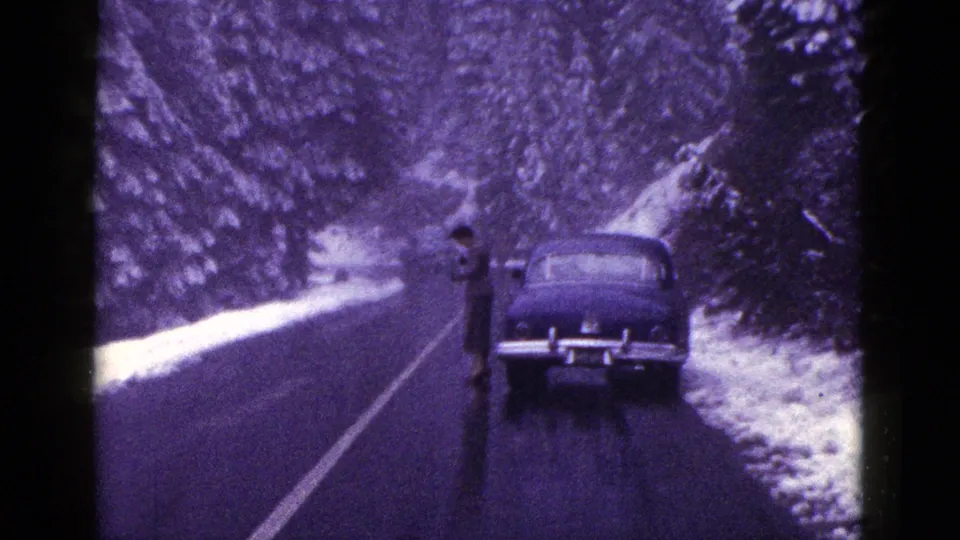 1953 California home-movie frame with an adult man standing beside a dark sedan on a wet snow-lined mountain road.