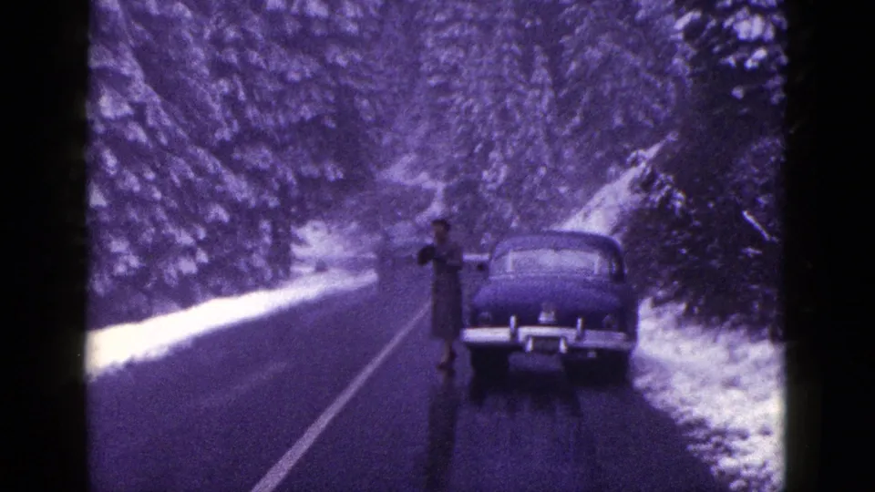 1953 California 8mm frame showing an adult beside a dark sedan on a wet road edged by snowbanks and trees.