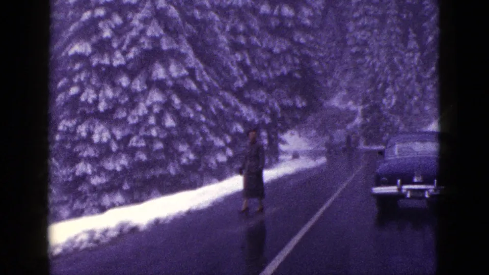 1953 California frame showing an adult man on a snow-lined mountain road while the sedan sits near the right edge.