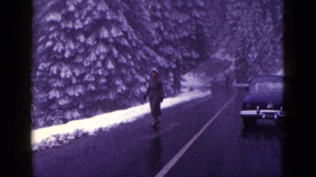 1953 California home-movie frame showing a woman on a wet snowy road with evergreen trees and a classic car nearby.
