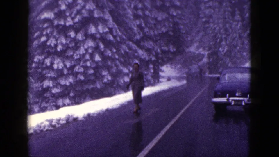 1953 California home-movie frame showing a woman on a wet snowy road with evergreen trees and a classic car nearby.