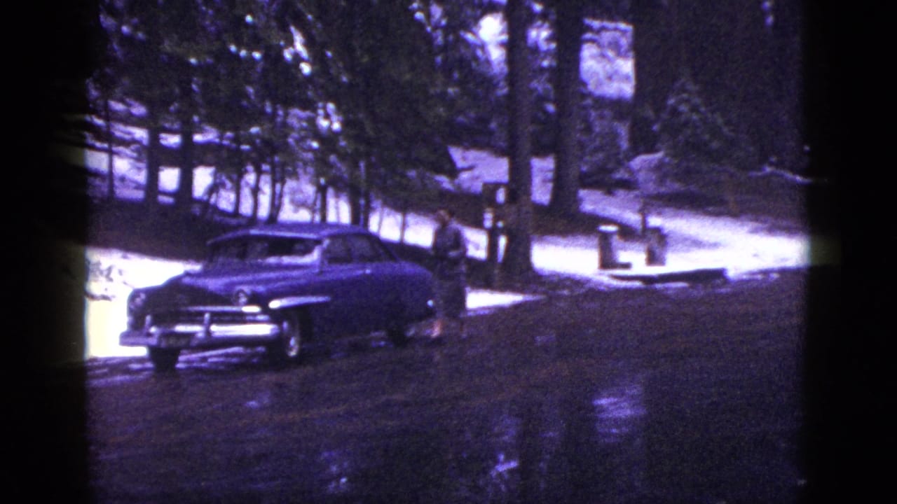 1953 California frame showing a dark mid-century sedan parked on a wet road among snowy forest and tall trees.