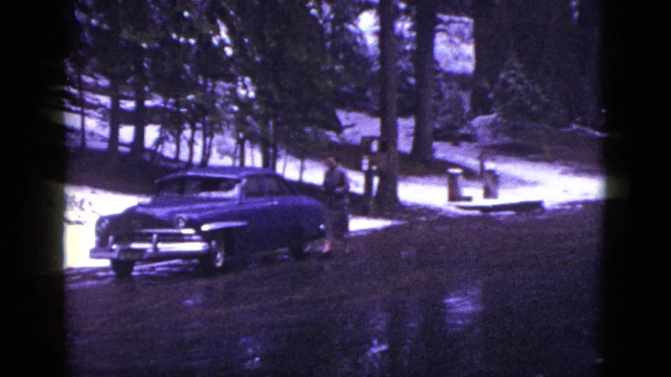 1953 California frame showing a dark mid-century sedan parked on a wet road among snowy forest and tall trees.