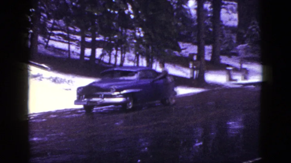 1953 California home-movie frame with a parked vintage sedan, snow patches, wet road, and dense redwood-like forest behind it.
