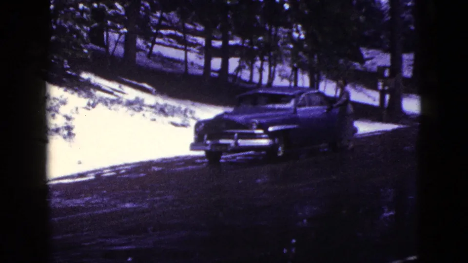 1953 California frame of a dark mid-century sedan parked beside snow and tall trees, rendered with blue-purple 8mm color shift.