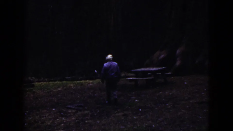 1953 California 8mm frame showing a man in a white cap walking toward a picnic table among large trees.