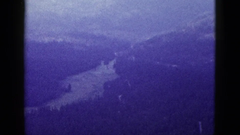 1953 8mm California aerial frame showing forested mountain slopes and a pale winding line through the valley floor.