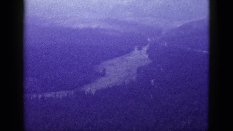 Aerial 8mm view of a hazy California mountain valley, with forested slopes and a pale river or road visible below.