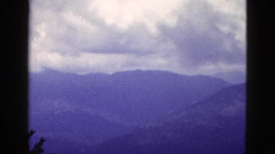 1953 California home-movie frame with layered mountain silhouettes, cloudy sky, and dark 8mm projection edges.
