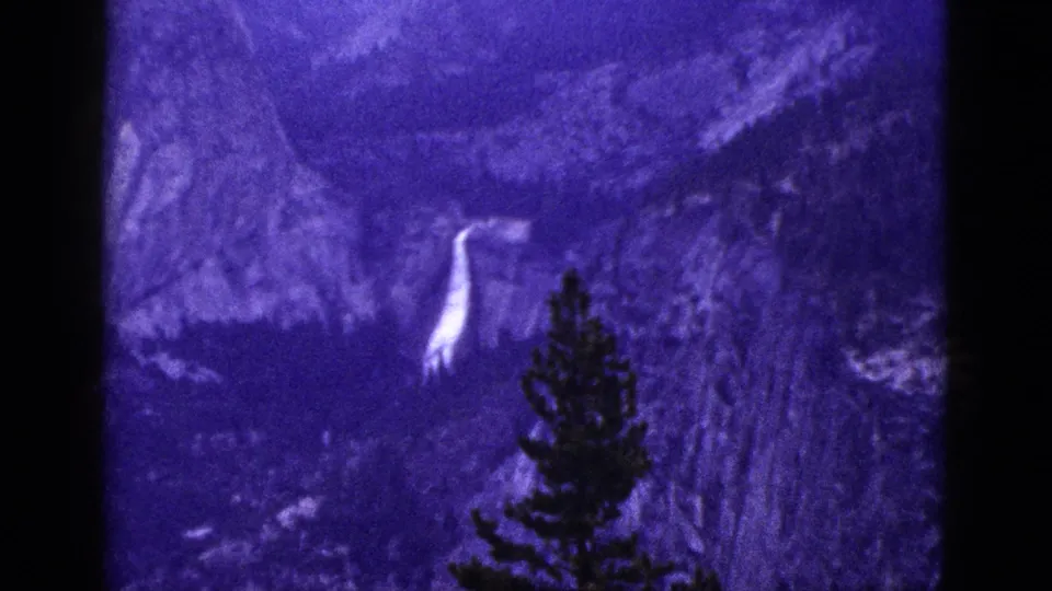 1953 California 8mm frame showing a rocky valley, bright waterfall, and dark pine tree silhouette in the foreground.