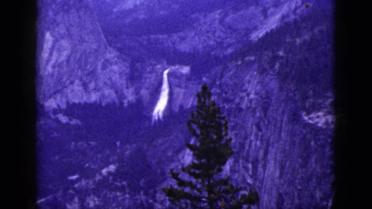 1953 California 8mm frame showing a white waterfall across a purple-toned mountain landscape with a pine silhouette in front.