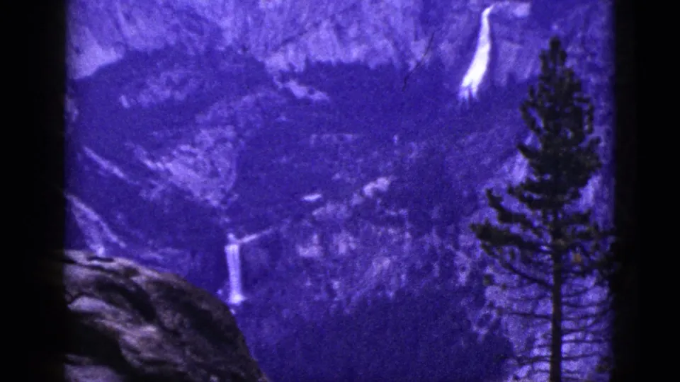 1953 California 8mm still with a distant waterfall, wooded slope, foreground pine, and visible dark frame borders.