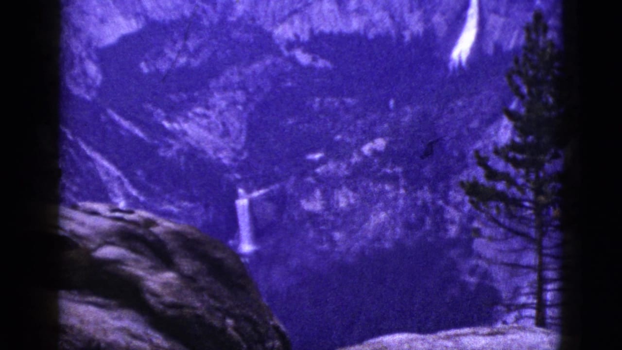 Purple-tinted 8mm frame of a distant waterfall between rocks and pine trees.