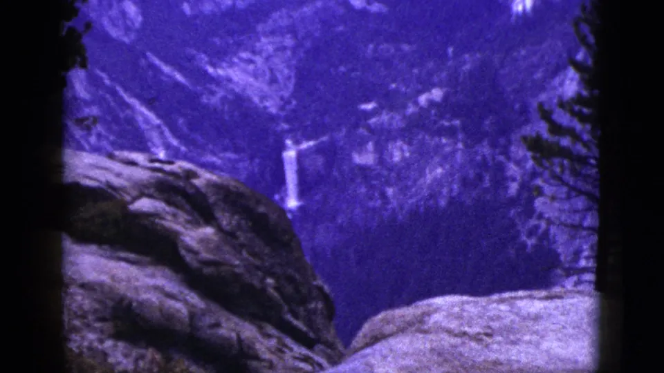 1953 California 8mm frame showing falling water on a forested slope, with dark rocks and pine trees cutting across the view.