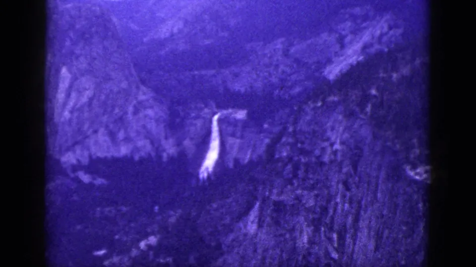 1953 California 8mm home-movie still with a narrow waterfall visible across a shadowed, wooded mountain slope and heavy film grain.