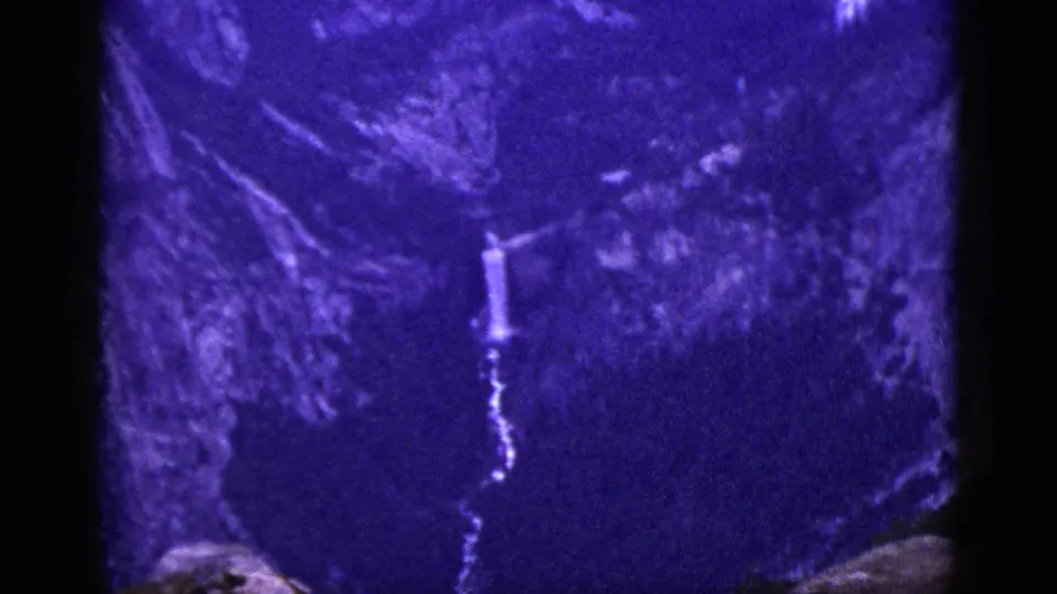 1953 California home-movie frame with a narrow waterfall visible on a rugged mountain face in purple-blue shifted color.