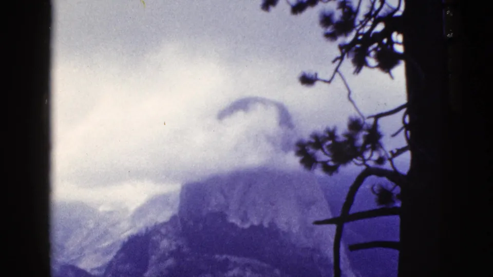1953 California home-movie frame with a cloud-wrapped Yosemite-like granite peak seen through dark pine branches.