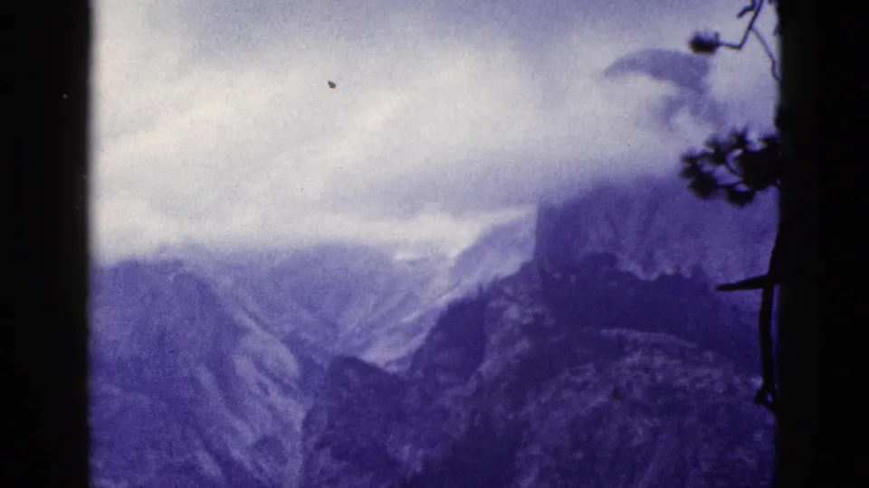 1953 California 8mm frame showing distant mountain ridges under cloud cover, with dark pine silhouettes at the edges.
