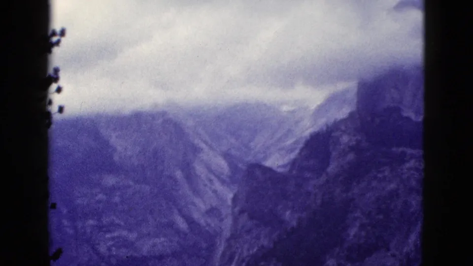 1953 California 8mm frame with a dark pine trunk in the foreground and cloudy mountain ridges beyond.