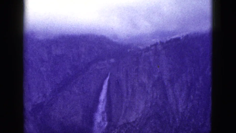 Purple-blue 1953 California 8mm frame showing a hazy mountain slope with a narrow pale waterfall-like streak below the cloudy sky.