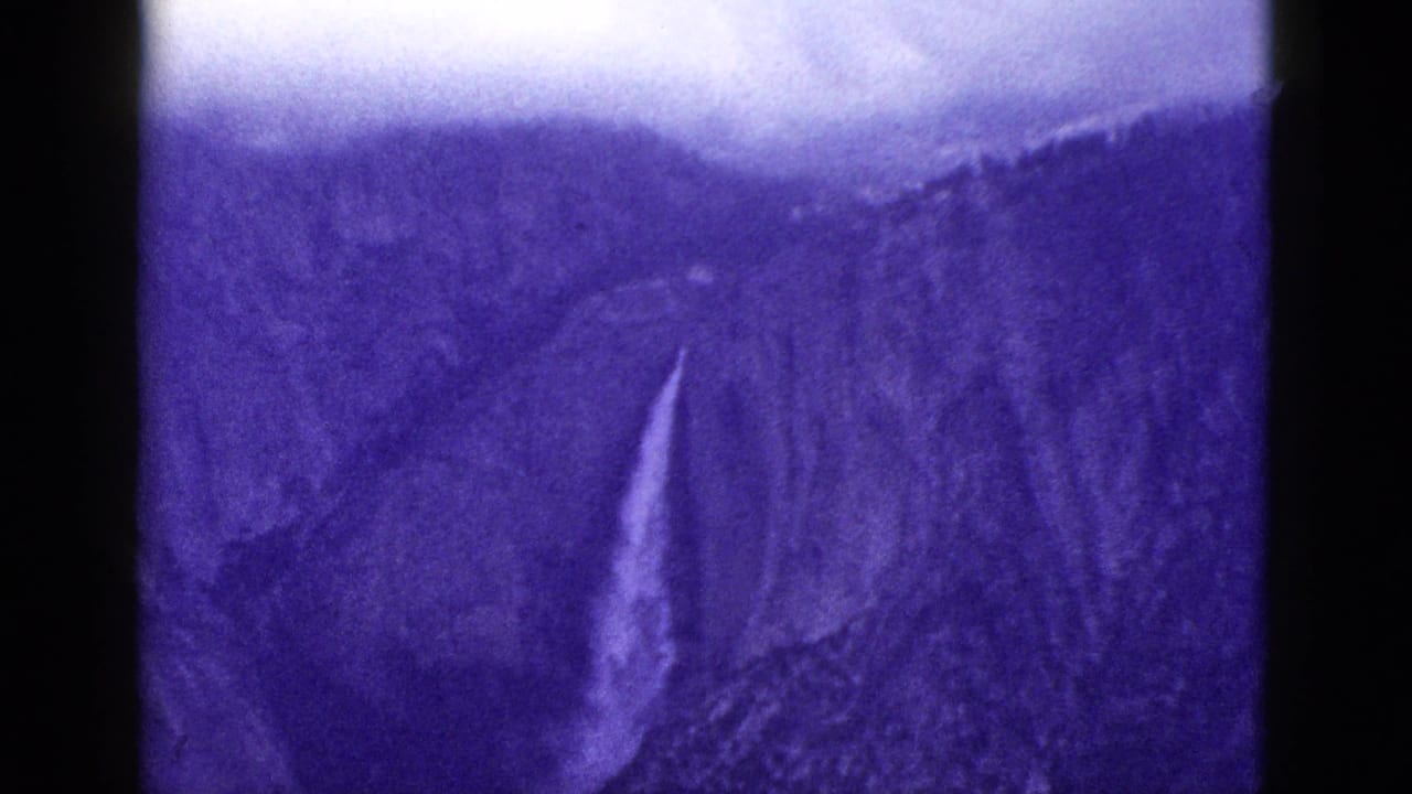 1953 California home-movie frame showing a tall waterfall descending a rugged, shadowed rock slope in purple-tinted 8mm footage.