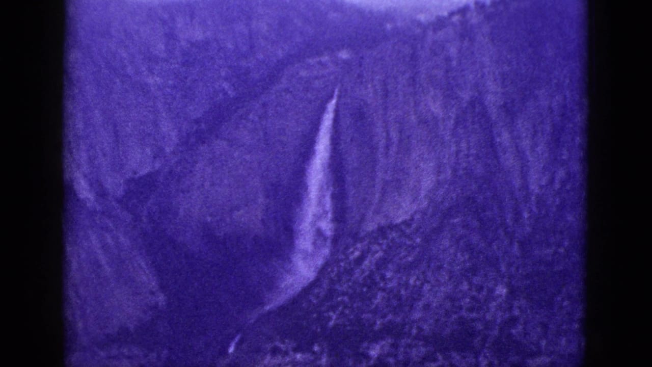 Purple-tinted view of a waterfall beside rugged mountain rock and shadowed terrain.