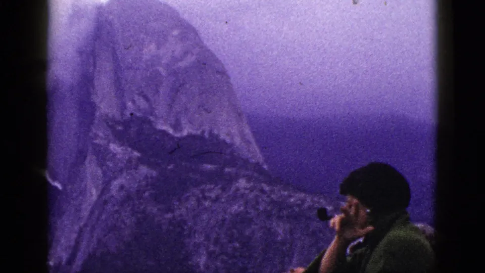 1953 8mm California footage shows a foreground man with a pipe near his mouth, framed against a hazy mountain peak and purple sky.