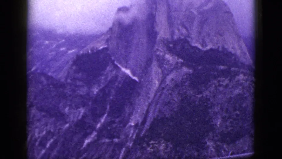 Color-shifted 1953 California mountain view with steep rock face, ridges, grain, and dark 8mm frame edges.