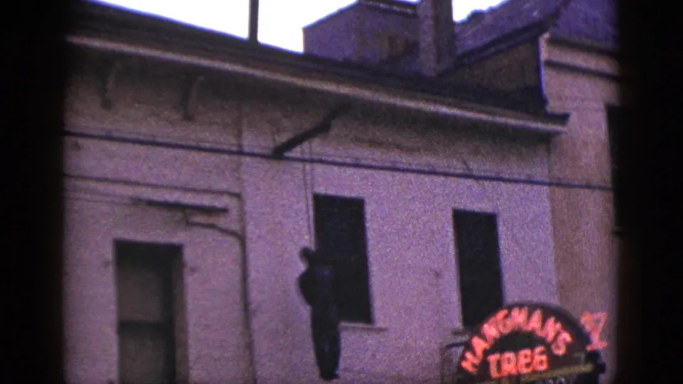 1953 California home-movie frame showing a historic-site building facade with dark window openings and a partial Hangman's Tree sign.