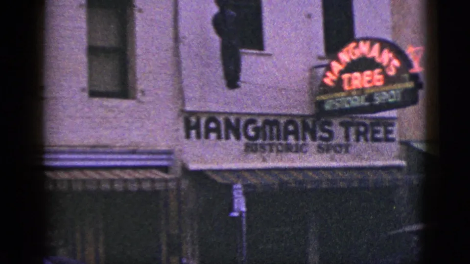 1953 California frame with Hangman's Tree Historic Spot signage, red neon, building windows, and a hanging mannequin-like figure.