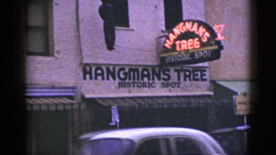 1953 California 8mm view of Hangman's Tree signage, red neon, awnings, a small street sign, and a passing car roof.