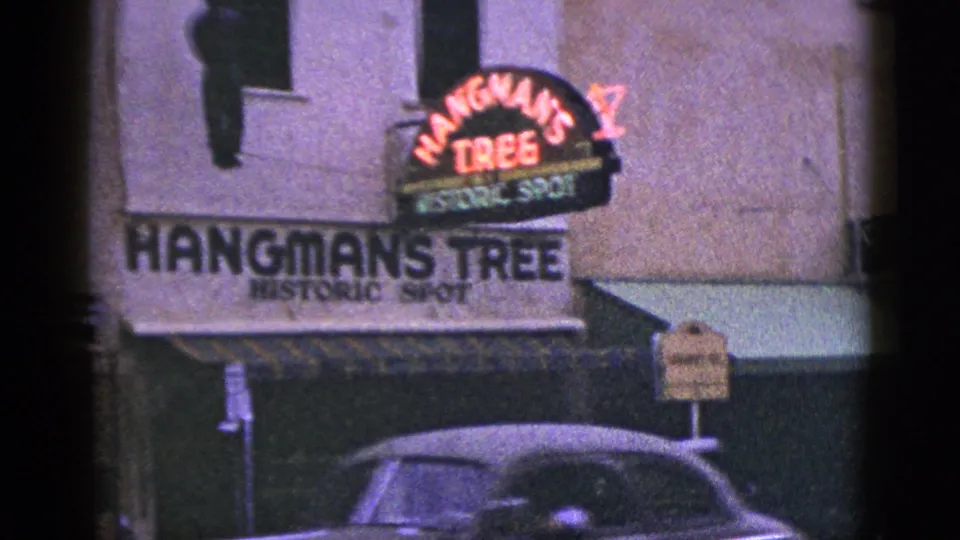 1953 California frame showing Hangman's Tree Historic Spot signage, neon lettering, awning, street sign, and a partial car below.