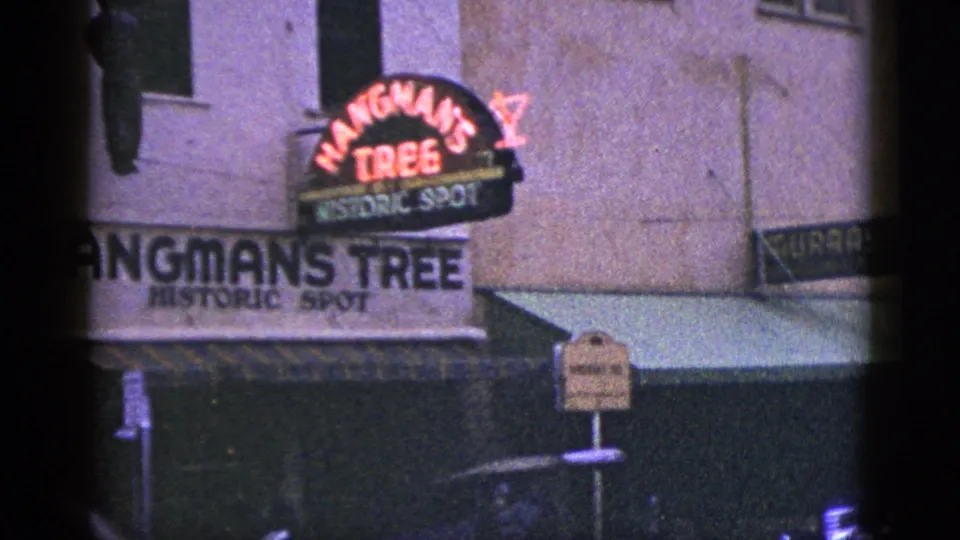 1953 California 8mm frame showing the Hangman's Tree historic spot sign on a storefront facade with awning and street signage.