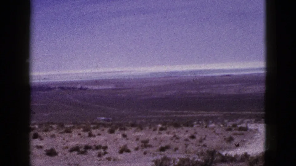 1953 California 8mm frame showing open desert terrain, scattered scrub, distant mountains, and a muted overcast sky.