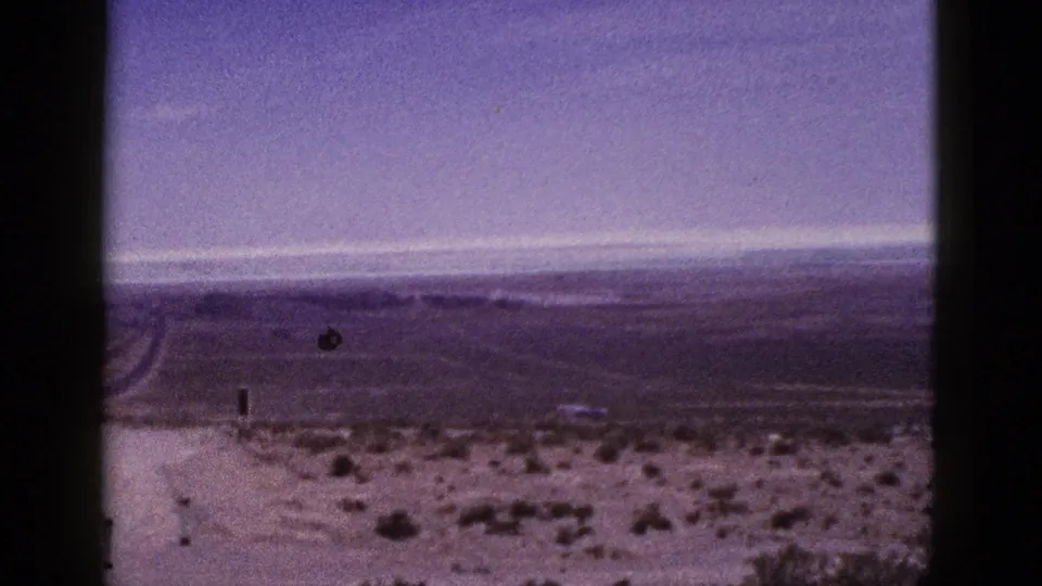1953 California home-movie frame with an empty desert road running into a broad scrub plain below distant hills and overcast sky.